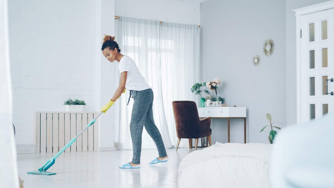 Jeune femme afro-américaine élégante en train de laver le sol chez elle avec une serpillière plate en plastique, portant des gants et une tenue confortable pour la maison. Intérieurs contemporains, propreté et jeunesse au cœur du concept.