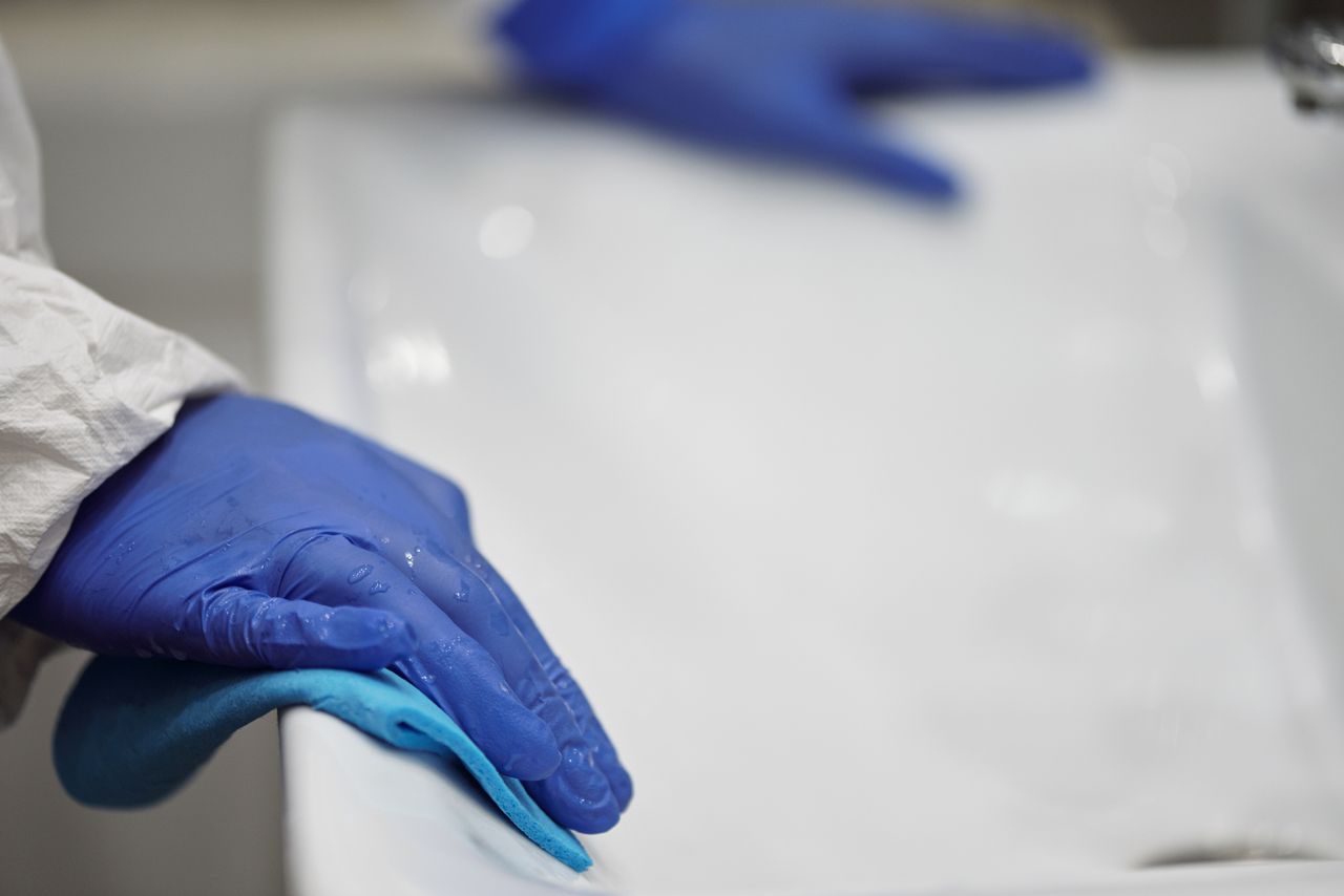 Gros plan sur une personne qui désinfecte un lavabo de salle de bain avec des gants et un chiffon, en mettant l’accent sur l’hygiène et la propreté.
